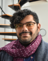 Photo of Sunny, a brown skinned person with curly black hair and a beard. He's wearing a scarf with IPA characters and a cardigan. In the background there is a spiral staircase.