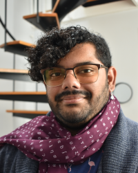 Photo of Sunny, a brown skinned person with curly black hair and a beard. He's wearing a scarf with IPA characters and a cardigan. In the background there is a spiral staircase.