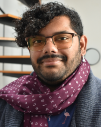 Sunny, a brown skinned person with curly black hair and a beard. He's wearing a scarf with IPA characters and a cardigan.