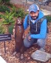 Rick posing with a metal statue of Otto the otter in a wooded area.