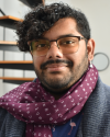 Sunny, a brown skinned person with curly black hair and a beard. He's wearing a scarf with IPA characters and a cardigan.